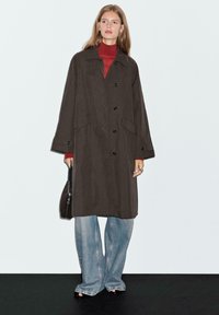 Dunkelbrauner langer Mantel mit großen vorderen Taschen, Knopfverschluss und spitzem Kragen, kombiniert mit einem roten Rollkragenpullover und weitem Jeans.