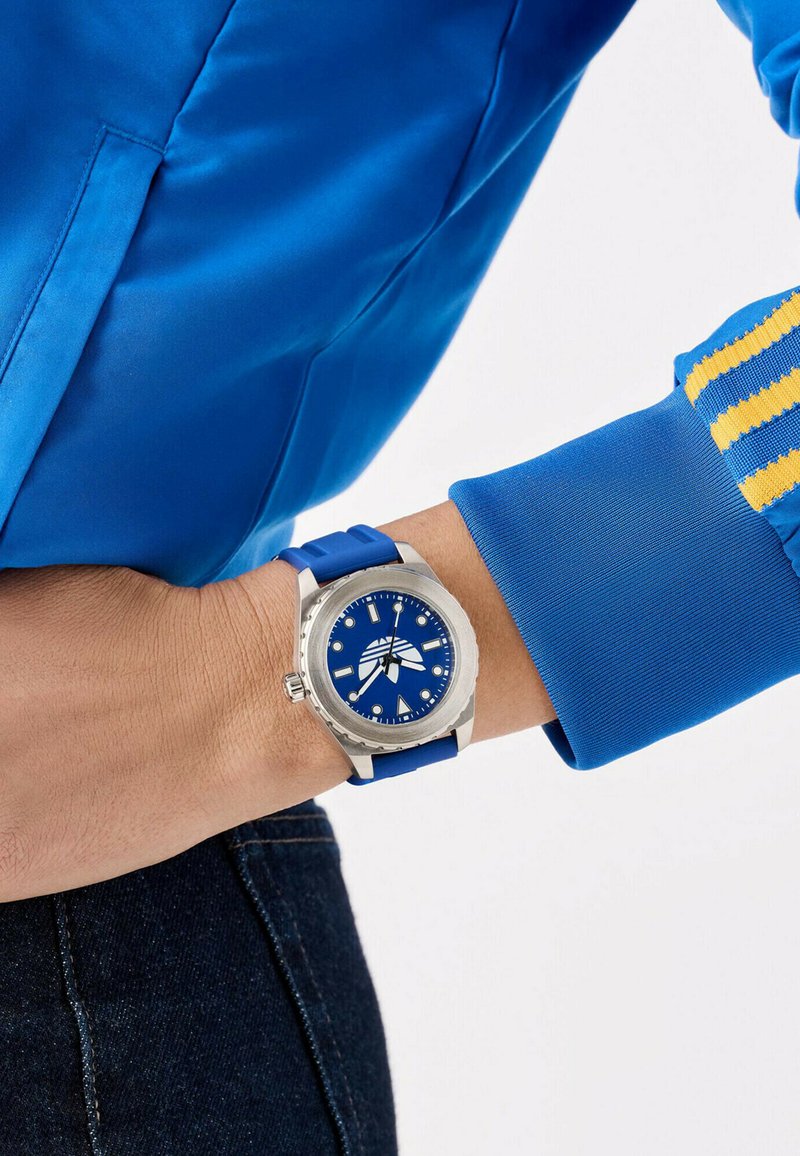 Modra ura s pasom iz nerjavečega jekla, modrim številčnikom in luminescentnimi kazalci, predstavljena na modrem silikonskem traku, v paru z modrim jopičem.