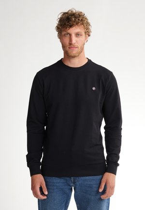 Sudadera negra de tela texturizada, con cuello redondo, mangas largas y un pequeño logo en el pecho. Llevada con jeans azules.