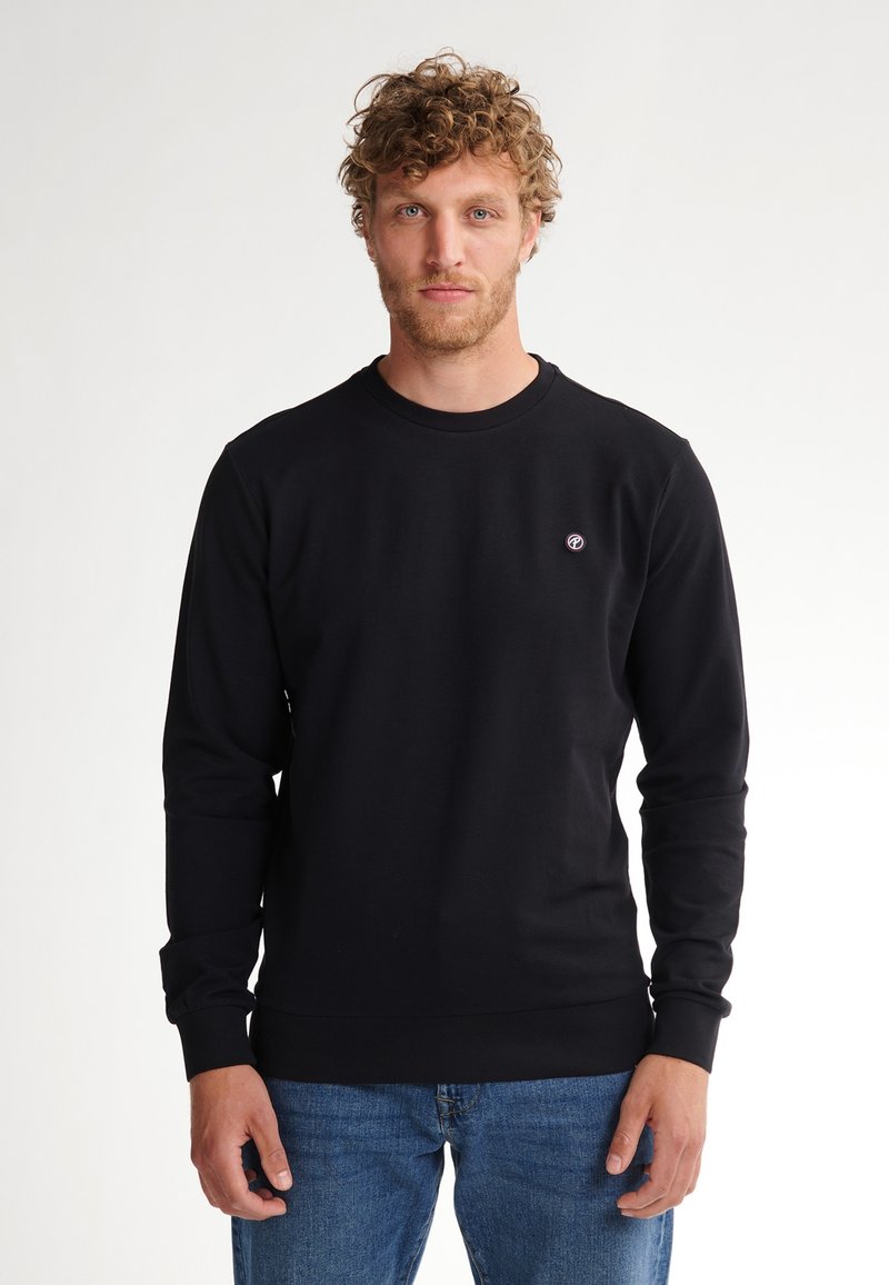 Sudadera negra de tela texturizada, con cuello redondo, mangas largas y un pequeño logo en el pecho. Llevada con jeans azules.
