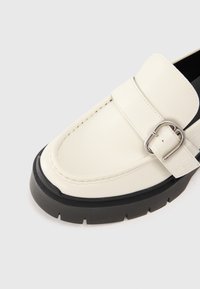 Valkoinen nahkainen loafer, jossa on paksu musta kumipohja. Kenkässä on koristeellinen hopeinen solki ja ompelut yksityiskohdat yläosassa.