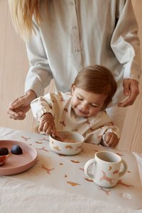 Suction bowl with cloud pattern, pastel colors; pink plate with fruit; white mug with handles; child in a printed bib, playful interaction.