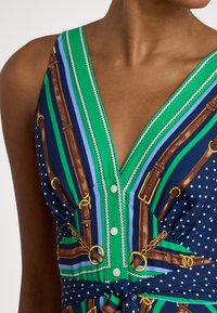 Κοντινό πλάνο αμάνικου φορέματος με πράσινες, μπλε και ναυτικές ρίγες, μοτίβα με άλογο και ζώνη, κουμπιά μπροστά και λεπτομέρεια δεμένης μέσης σε σκουρόχρωμο δέρμα.