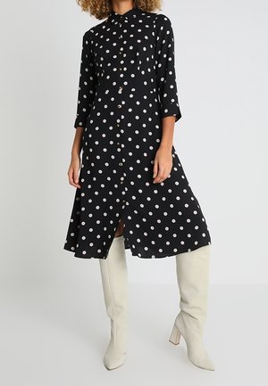 Vrouw in een zwarte midi hemdjurk met witte stippen en witte kniehoge haklaarzen, staande tegen een effen achtergrond.