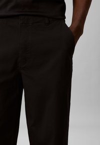 Άνδρας που φοράει μαύρο παντελόνι με το χέρι χαλαρά τοποθετημένο στην εμπρόσθια δεξιά τσέπη, σε ένα μονόχρωμο γκρι φόντο.