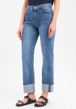 Persoon die blauwe omgeslagen spijkerbroek en zwarte sandalen met gesp draagt, staand tegen een witte achtergrond.