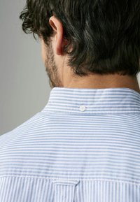 Chemise rayée bleu clair et blanche avec un col court, fermeture à boutons au dos, et un tissu texturé pour plus de détails.