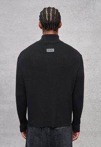 Schwarzer Rippstrickpullover mit hohem Kragen und einem kleinen Etikett auf der Rückseite. Verfügt über enganliegende lange Ärmel und ein leicht verkürztes Design.