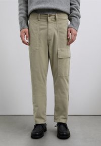 Pantalones cargo de color beige claro con bolsillos laterales, cierre frontal con botones y ajuste de pierna recta, combinados con zapatos negros y un suéter gris.