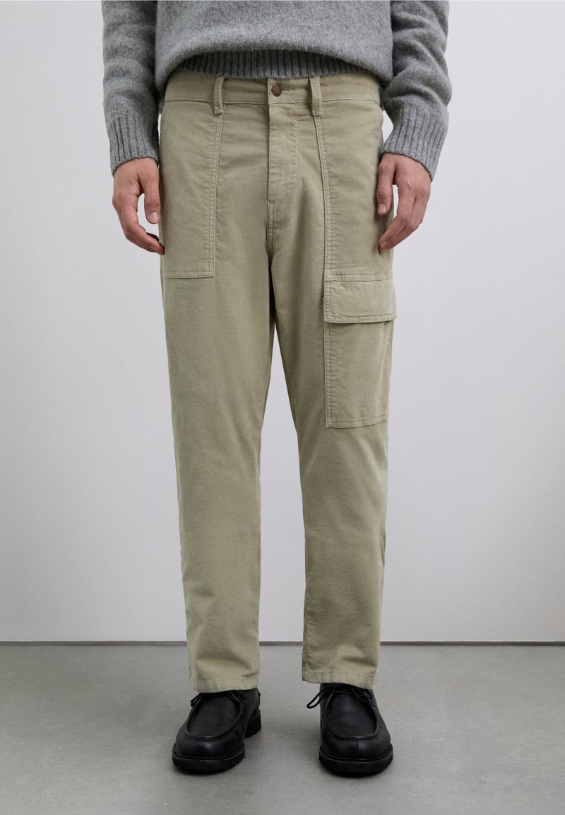 Pantalones cargo de color beige claro con bolsillos laterales, cierre frontal con botones y ajuste de pierna recta, combinados con zapatos negros y un suéter gris.