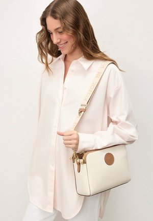 Femeie îmbrăcată într-o cămașă roz deschis cu nasturi, purtând o geantă crossbody crem texturată cu accente maro deschis și accesorii aurii.
