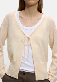 Brzoskwiniowy kardigan o miękkim, lekkim splocie, z otwartym przodem i związaniem z przodu. Posiada długie rękawy i ściągacze u mankietów.