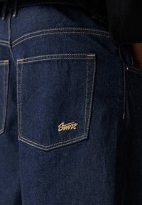 Jean en denim bleu foncé avec surpiqûres contrastantes et poches arrière. Un logo brodé beige est visible sur la poche droite.
