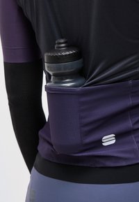 Ποδηλάτης που φοράει μωβ φανέλα με ένα μαύρο παγούρι νερού κολλημένο στην πίσω τσέπη της φανέλας.