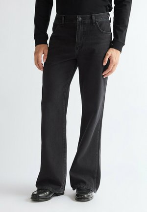 Persona che indossa jeans neri a gamba larga, top nero a maniche lunghe e scarpe di pelle nere, in piedi davanti a uno sfondo chiaro e uniforme.