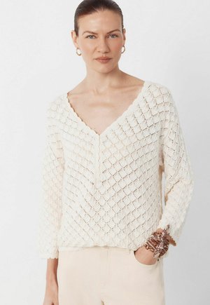 Žena se zlatými kruhovými náušnicemi nosí krémový texturovaný svetr s výstřihem do V a světlé kalhoty, s hnědým vzorovaným šátkem uvázaným kolem zápěstí.