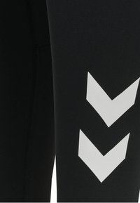 Svarta leggings med en slät textur, kroppsnära design och djärva vita chevronmönster på sidan, med synliga sömnadsdetaljer.