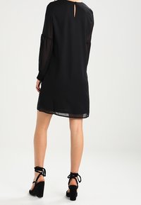 Schwarzes Kleid mit durchsichtigen langen Ärmeln, lockerer Passform und einem Schlüsselaugenverschluss hinten. Kombiniert mit schwarzen Sandalen mit Absatz und Knöchellaschen.