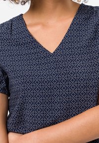 Primer plano de una persona que lleva una blusa de cuello en V de color azul marino con un patrón y mangas cortas, con los brazos cruzados a la altura de la cintura.