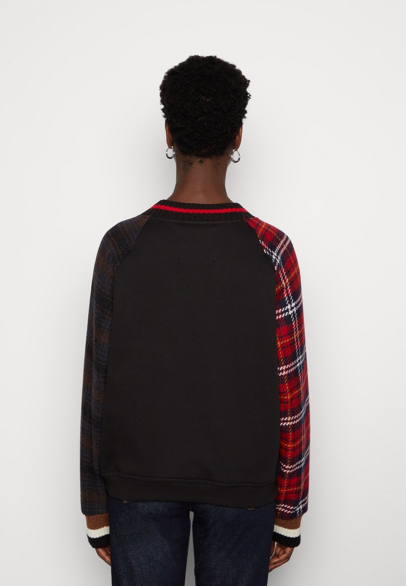 Osoba z krótkimi, kręconymi włosami ubrana w czarną bluzę z mismatched rękawami w kratę, jeden czerwony, a drugi ciemny, stojąca na tle białym.