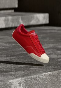 Κόκκινο αθλητικό παπούτσι Converse με λευκό κάλυμμα δακτύλων, στον αέρα πάνω από σκάλες από τσιμέντο, ρίχνοντας σκιά στο έδαφος.