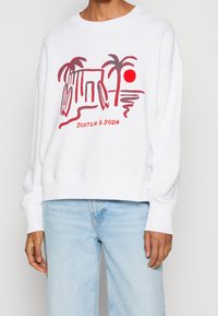White sweatshirt with a relaxed fit features a red graphic of palm trees and a sun, labeled "SCOTCH & SODA" at the bottom.
