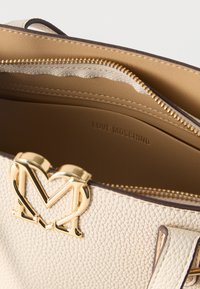 Τσάντα χειρός από δέρμα με υφή κρέμας, με κούμπωμα με λογότυπο M σε σχήμα χρυσής καρδιάς και εσωτερική τσέπη με φερμουάρ σε ταμπά χρώμα που φέρει ανάγλυφη την επιγραφή «Love Moschino».