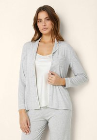 Grå pyjamasuppsättning som inkluderar en knapp-up, långärmad topp med ficka och kontrasterande piping, tillsammans med en vit spets och matchande byxor.