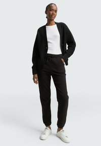 Schwarze gerippte Strickjacke über einem weißen Oberteil, kombiniert mit schwarzen Jogginghosen und weißen Turnschuhen. Die Strickjacke hat große Knöpfe und eine lockere Passform.