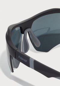 Mustad mustasportide prillid hallide aktsentidega, millel on voolujooneline disain, tumedad objektiivid ja kummised nina padjad mugavuse tagamiseks. Polaroidi logo on nähtav.