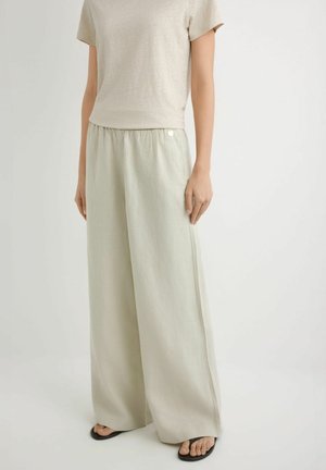 Kobieta w luźnych, jasnobeżowych spodniach o szerokich nogawkach z elastycznym pasem i beżowej bluzce z krótkim rękawem, stojąca na jednolitym tle.