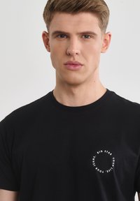 Černé bavlněné tričko s kruhovým bílým logem na levé části hrudi. Design s kruhovým výstřihem a krátkými rukávy. Hladká textura.