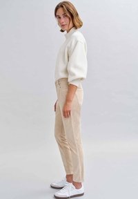 Beige Cord-Hosen mit schlanker Passform, kombiniert mit einem weißen Strickpullover und weißen Sneakern. Der Pullover hat einen hohen Kragen und kurze Ärmel.