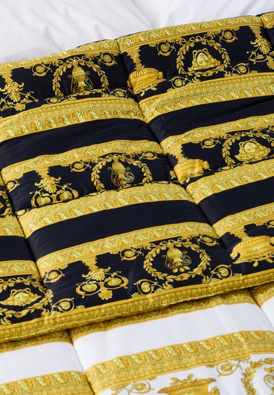 Couverture avec un motif à rayures dorées et noires, des motifs complexes et une texture satinée, posée sur une housse de couette blanche.