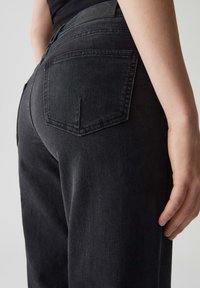 Jean en denim noir vu de l'arrière, avec deux poches arrière rectangulaires et une texture lisse, présentant un léger éclat.