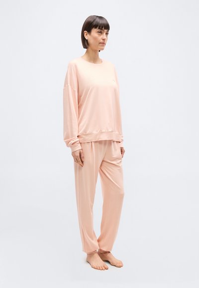 Γυναίκα που στέκεται ξυπόλητη, φοράει φαρδιά, ανοιχτό ροζ μπλούζα με μακριά μανίκια και ταιριαστό παντελόνι μπροστά σε απλό ανοιχτό φόντο.