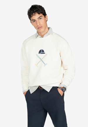 Sudadera crema con una gorra azul marino y mazos cruzados bordados en colores pastel. Combinada con pantalones azul marino y una camisa a rayas debajo.