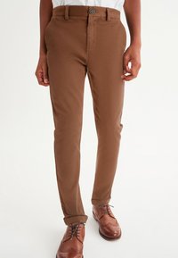 Pantalon marron ajusté à la coupe slim avec une texture lisse, doté d'une fermeture à boutons et de poches latérales, assorti à des chaussures habillées marron.