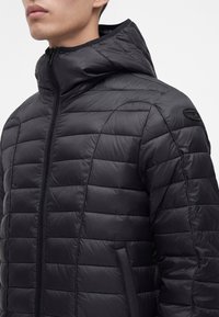 Homme portant une veste matelassée noire à capuche avec une fermeture éclair devant, des poches latérales et un petit écusson logo sur la manche supérieure.