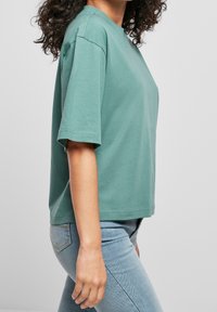 Krátký rukáv, tyrkysově zelené bavlněné tričko s uvolněným střihem, kulatým výstřihem a hladkou texturou, spárované se světle modrými džíny.