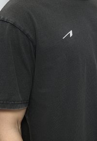 Κοντινή λήψη μαύρου κοντομάνικου μπλουζιού με μικρό λευκό γωνιακό λογότυπο στο στήθος, που φοράει ένα άτομο με ανοιχτόχρωμο δέρμα.