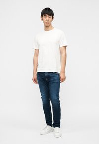 Hvid bomuld T-shirt, blå denimjeans og hvide sneakers. Pasformen er slank med et afslappet design. Baggrunden er ensfarvet hvid.