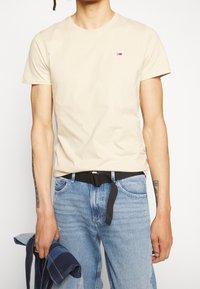 Beige bomulls t-shirt med rund halsringning och korta ärmar, med en liten logotyp på vänster sida. Bärs med blå jeans.