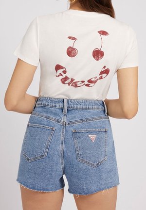 Femme portant un t-shirt blanc avec des cerises rouges et le texte "Guess" au dos, assorti à un short en denim bleu taille haute avec des poches arrière.
