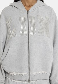 Ανοιχτό γκρι φούτερ με φερμουάρ και μεταλλικά νύχια που σχηματίζουν τη λέξη "PINK" στο στήθος και ξεφτισμένες άκρες στο στρίφωμα και στα μανίκια.