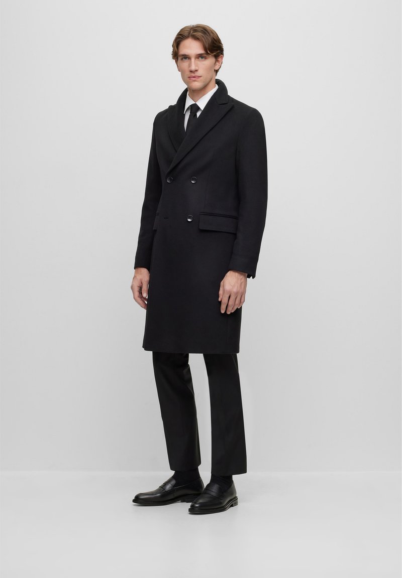 Manteau en laine noir à double boutonnage, avec un revers en pointe, des poches latérales et une coupe au genou, associé à un pantalon formel noir et des chaussures.