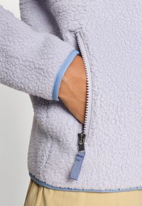 Lichtpaarse fleecetrui met een gestructureerd oppervlak; beschikt over een blauwe rits en cuffaccenten; zijzak zichtbaar aan de linkerkant.
