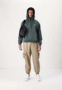 Mlad moški z dreadlocksi, oblečen v zeleni kapuco, bež cargo hlače, bele superge in črno torbico čez ramo, na enobarvni beli ozadju.