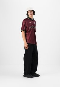 Bordo marškiniai su juodu apykakle ir geltonomis detalėmis, su logotipu ant krūtinės. Derinama su juodomis plačiomis kelnėmis ir juodais sportiniais bateliais.