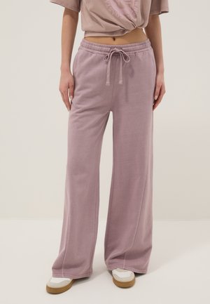 Personne portant un pantalon de survêtement ample lavande avec cordon, des baskets blanches et un t-shirt court taupe, debout devant un fond uni.